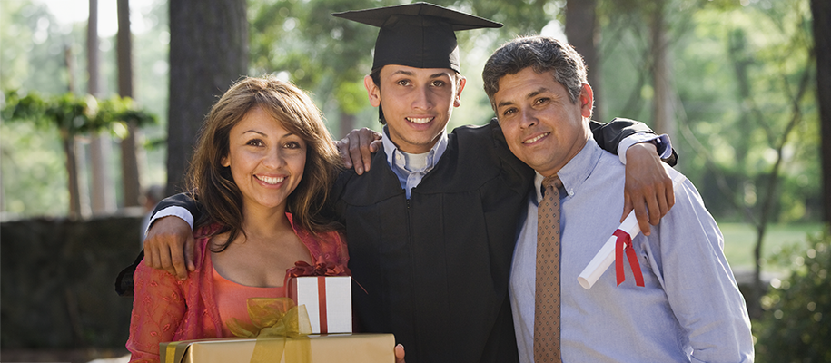 Latino graduate in black cap, gown with arms around parents, mom holding gifts