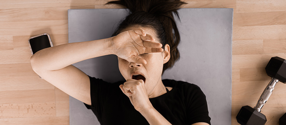 Asian woman in black T-shirt lying down on yoga mat, yawning, covering eyes