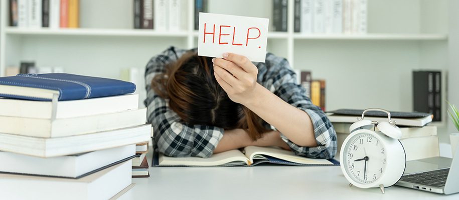 Asian student with head down on open book in library holding up sign saying Help