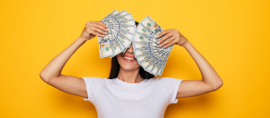 White woman in white shirt holding fans of cash over eyes on yellow backdrop