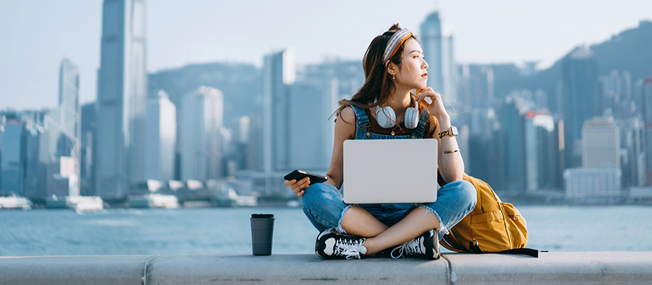 Asian woman in overalls in front of city skyline with laptop, contemplative