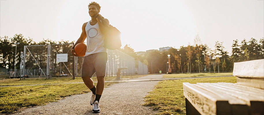 Black man in workout clothes carrying a basketball and gym bag walking on campus