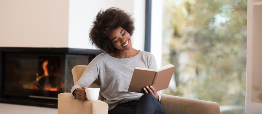 Black female w/ short curly hair holding coffee cup, smiling at book in armchair