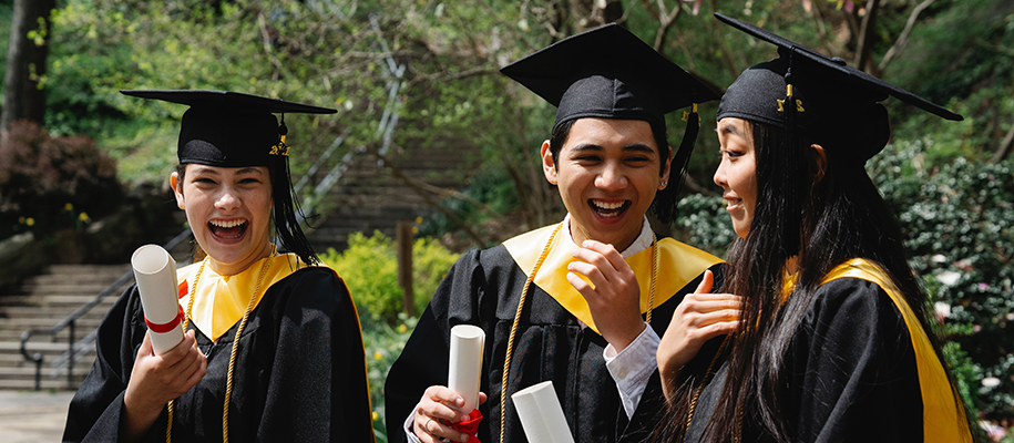 Three Asian students laughing in black and yellow graduation robes with diplomas