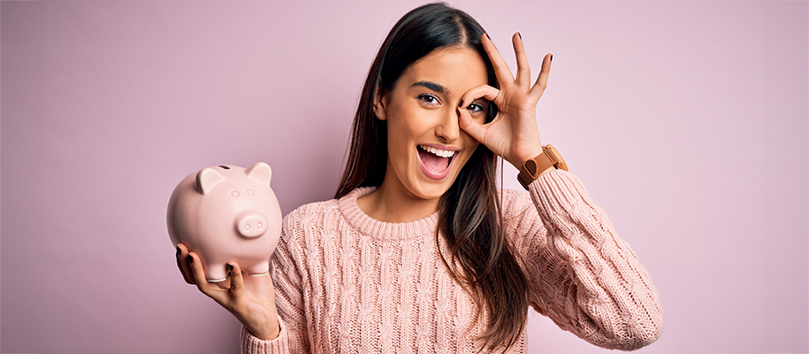 Middle-eastern woman in pink sweater holding piggy bank, okay hand sign to eye