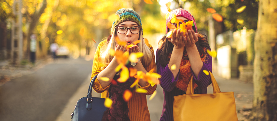 Two women in bright clothes, hats, with purses and glasses, throwing fall leaves