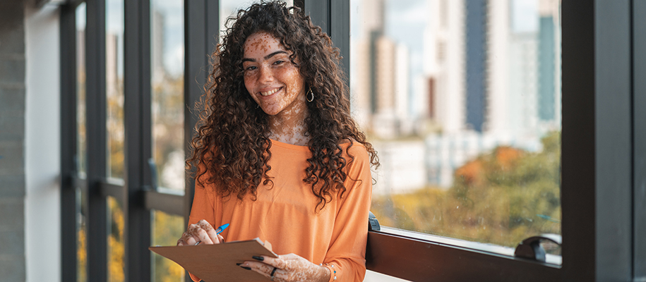 Black woman with curly hair, vitiligo in orange shirt with clipboard by window