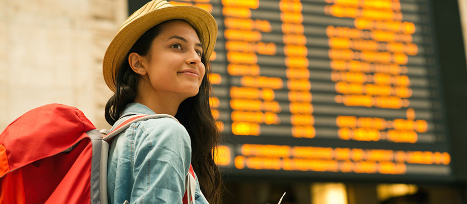 Woman with hat and red backpack smiling in front of boarding board
