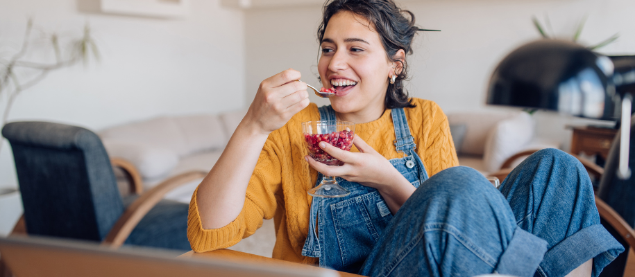 White female student in overalls eating bowl of pomegranate arils at desk