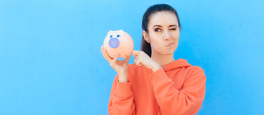 Black-haired female winking one eye thinking, holding and pointing at piggy bank