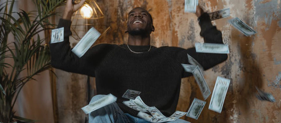 Black man sitting with hands in air and hundred-dollar bills flying around him