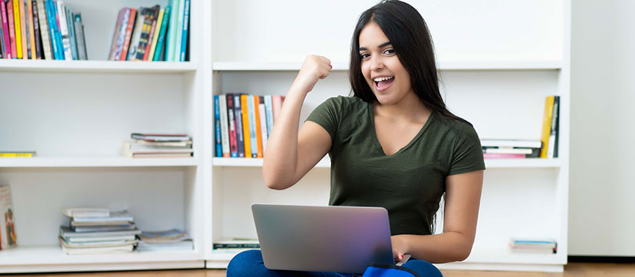 Happy Latina high school student holding laptop and raising fist in victory