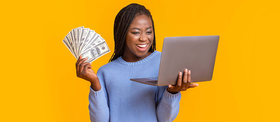 Young Black woman in blue shirt smiling, holding laptop and fan of $100 bills