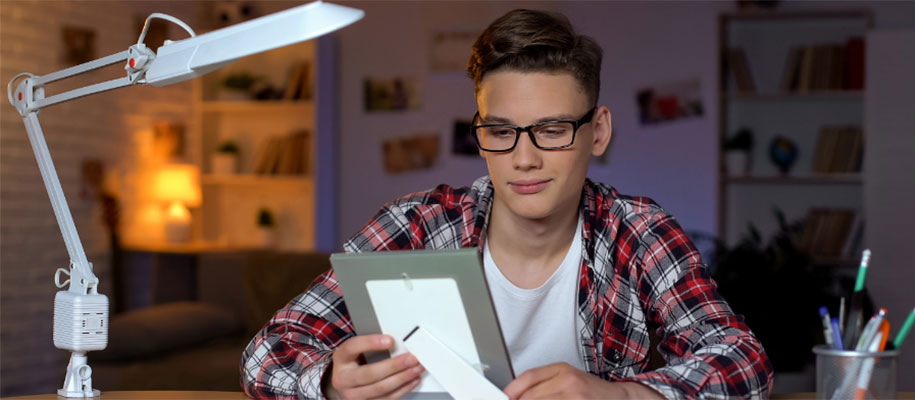 White male with glasses in dorm, holding & looking at framed photo at desk