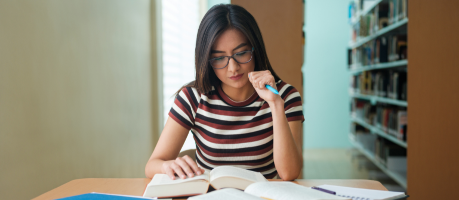 Female student in glasses, striped shirt studying in library with books and pen