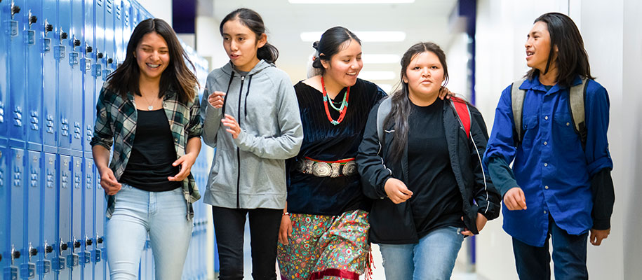 Five Native American teenage students walking down hall with lockers at school