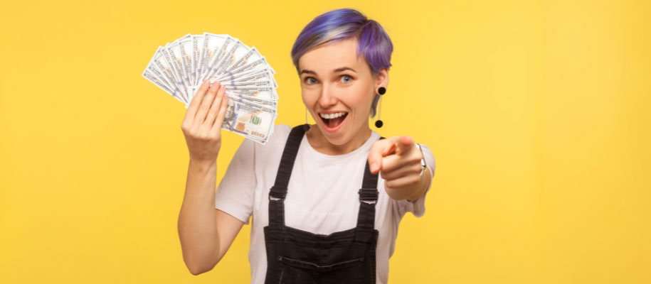 White woman with purple hair and overalls holding fan of $100 bills and pointing