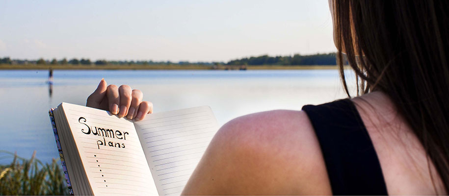 Black-haired female by lake holding notebook reading Summer Plans with bullets