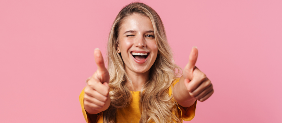 White blonde student winking her eye, giving 2 thumbs up against pink background