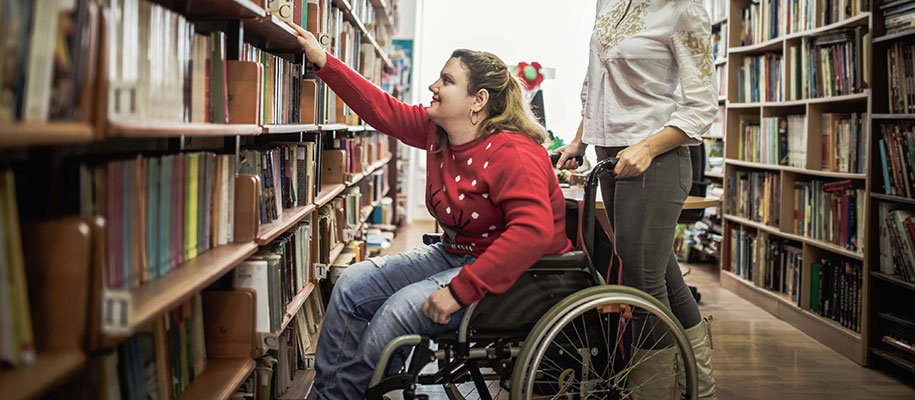 Blonde female in red sweater and wheelchair reaching for book in the library