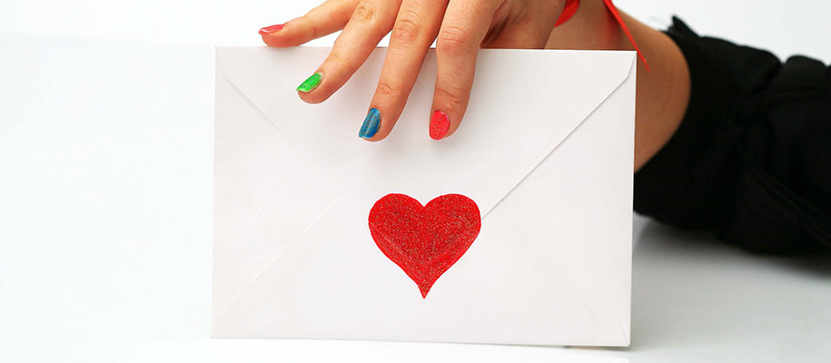 White hand with different colored nails holding white envelope with red heart