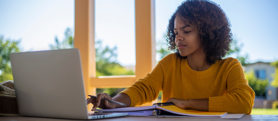 Young Black woman in yellow sweater on computer with open binder