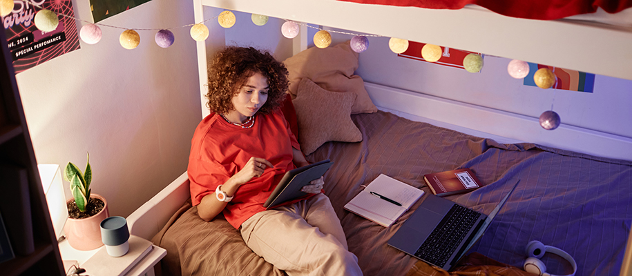 Woman with curly hair in orange shirt reclined on bed w/ notebook, book, tablet