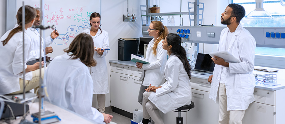 Five diverse students in lab listening to White female, Black male professors