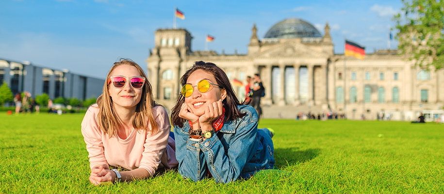 Two women with sunglasses lying in grass outside building with German flags