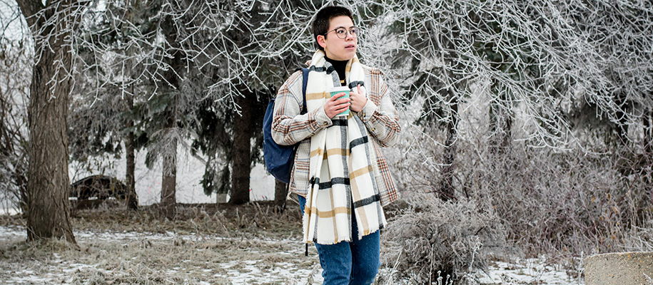 Non-binary multiracial person in scarf and winter coat, holding coffee and walking on campus in winter