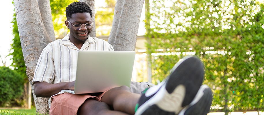 Black college student in striped shirt and glasses, sitting and leaning against tree with his laptop