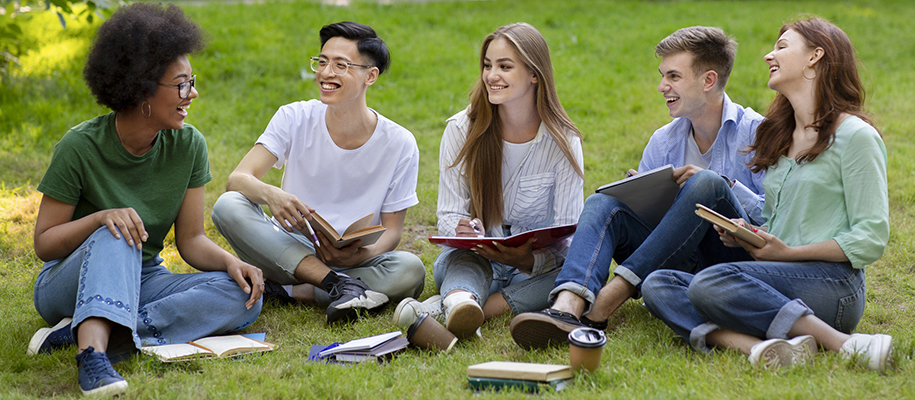 Diverse group of five students sitting on grass with books and pens, laughing