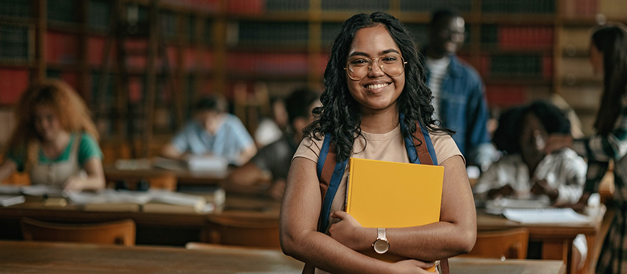 Indian woman in glasses hugging book, smiling with library of students behind