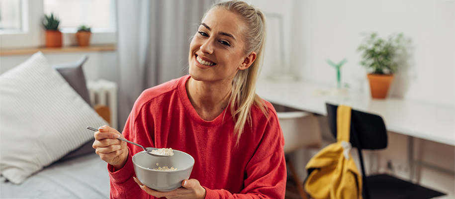 Smiling White woman in orange shirt sitting on dorm bed with bowl of oatmeal