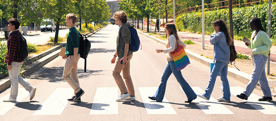 Group of six diverse students walking across crosswalk with books, bags