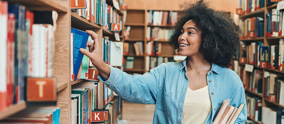 Smiling Black woman in library holding books, perusing and touching bookshelves