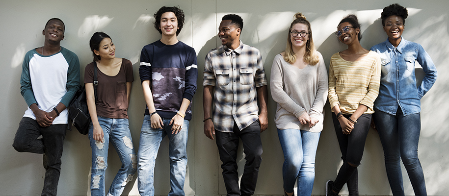 Seven diverse students standing against wall outside, smiling at each other