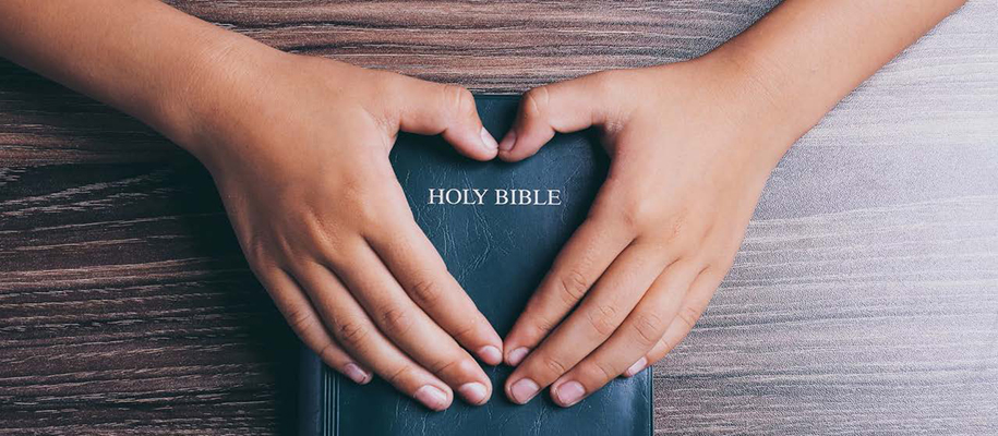 Hands making heart over cover of Holy Bible on wooden table