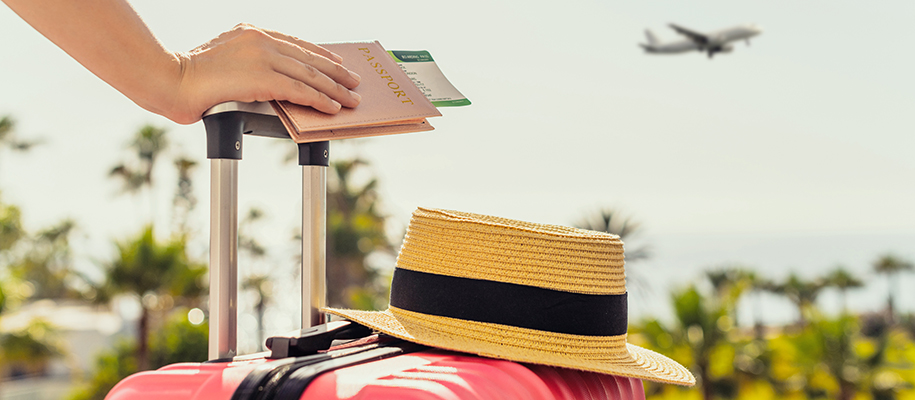Hand holding passport and plane tickets on handle of suitcase with hat