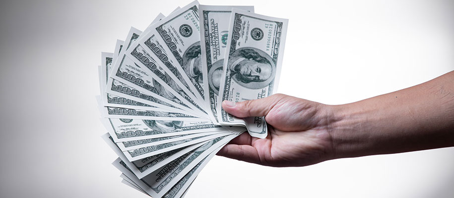 White hand holding multiple fanned out $100 bills on white backdrop