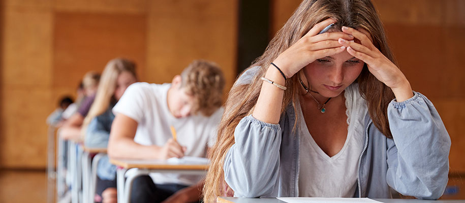 Line of students taking tests at desks with focus on White girl holding forehead