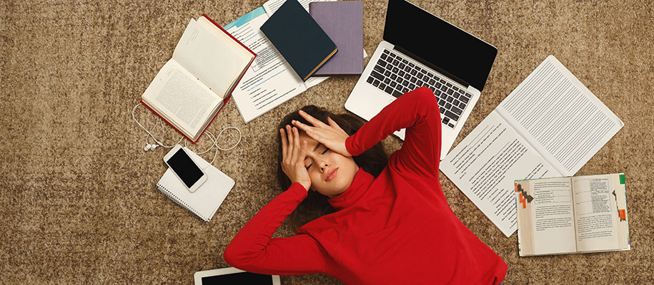 Tired female student lying on floor by books and gadgets with hands on her head
