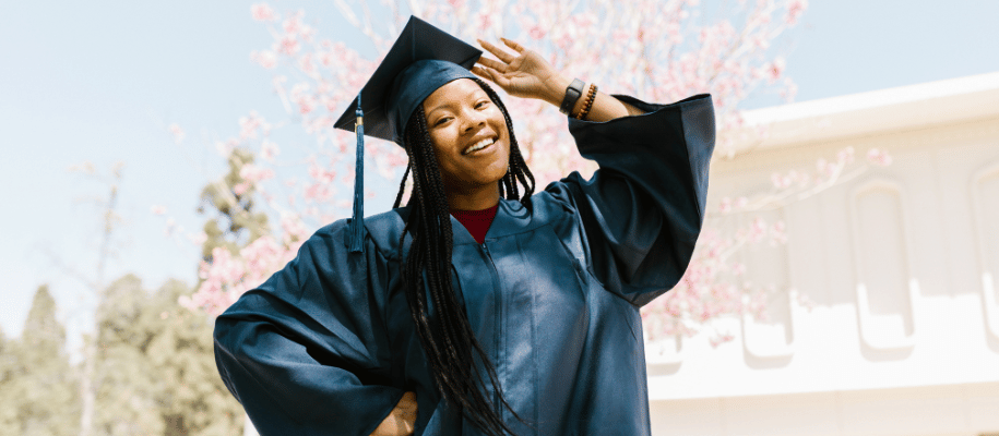 Group of graduates in dark green gowns throwing caps in air on blue sky day