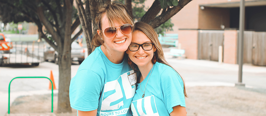 Two blonde women wearing blue community service shirts leaning into each other