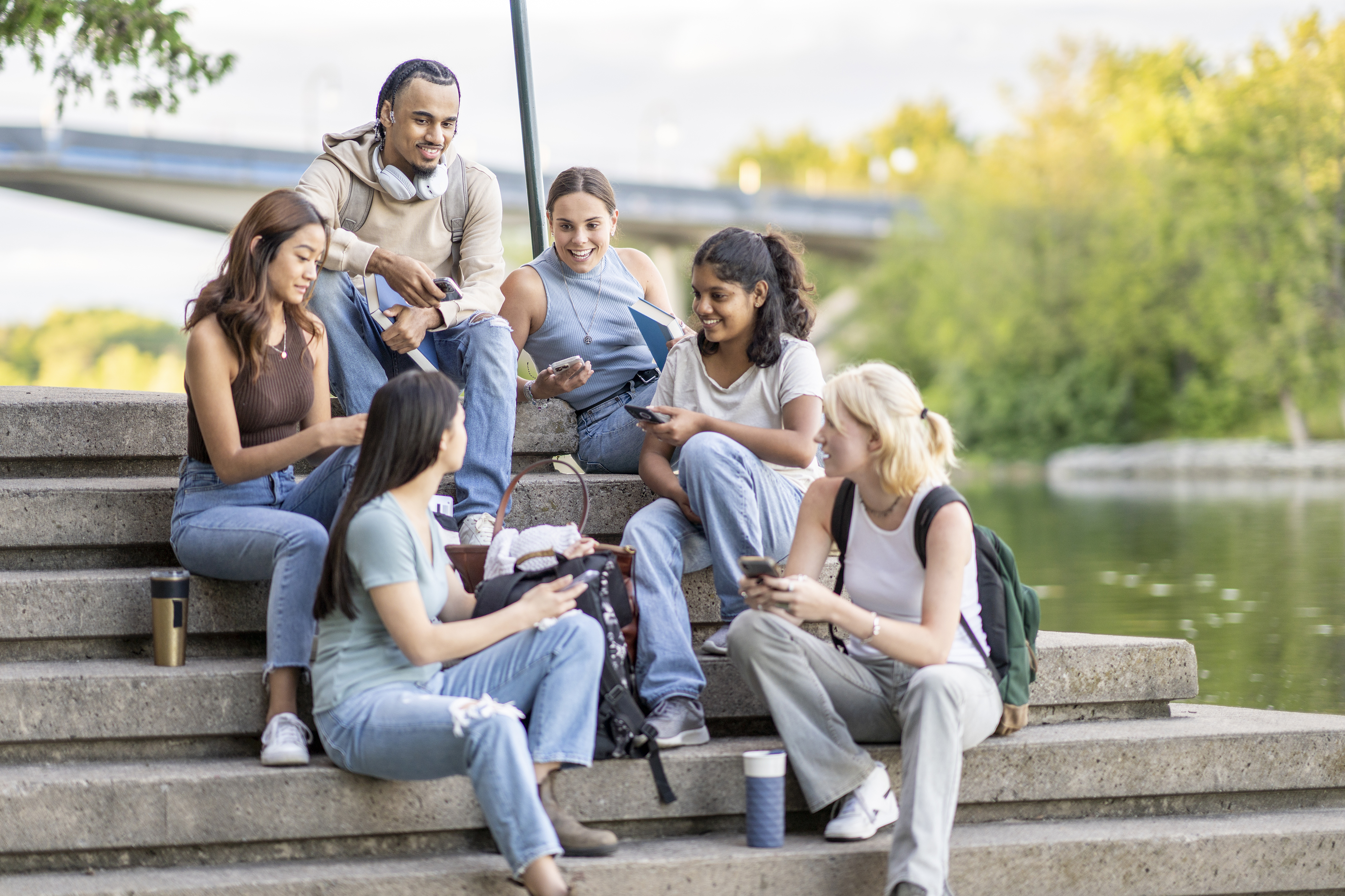 Group of diverse students in casual clothes sitting on stairs with phones, coffee