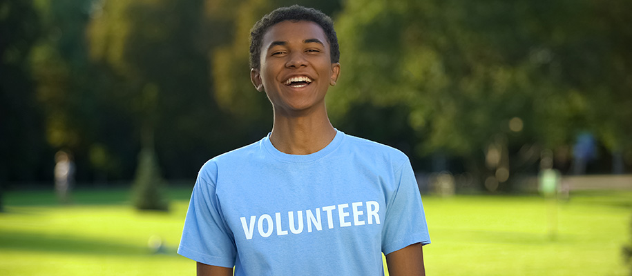 Young Black male teen smiling in light blue volunteer shirt outside in park