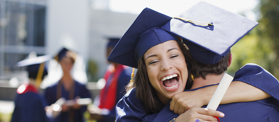 Brunette girl in blue grad cap and gown, smiling with arms around neck of peer