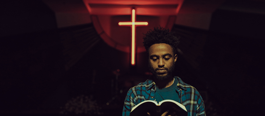 Black man in blue plaid shirt holding book with red lit cross behind him