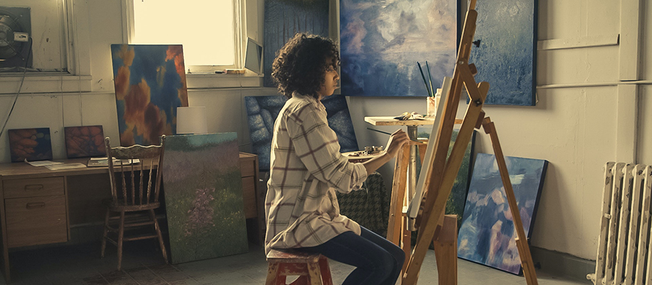 Black female artist with short curly hair in art studio, painting at an easel