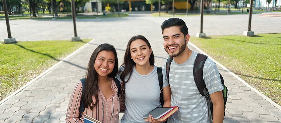 Two Hispanic women and one man smiling on campus with backpacks and books